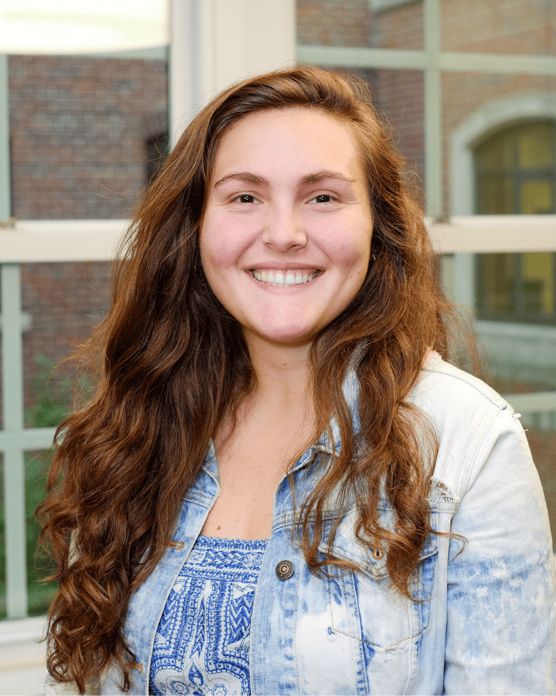Wellness Center staff member smiling in a portrait, standing indoors in front of a window with a brick building visible outside.